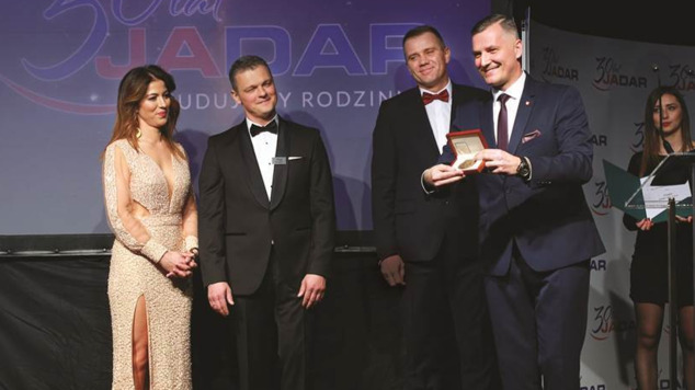Wicemarszałek Rafał Rajkowski wręcza medal przedstawicielom firmy Jadar podczas gali. Kobieta w wieczorowej sukni oraz trzej mężczyźni w strojach oficjalnych stoją na scenie. W tle kobieta prowadząca uroczystość.