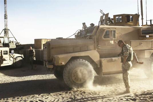 Żołnierz biorący udział w misji w Afganistanie stoi przy pojeździe wojskowym. W tle koleny pojazd i żołnierz stojący przy metalowym maszcie.