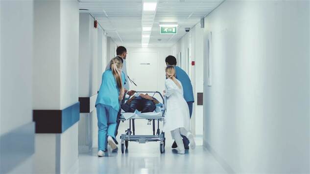 Personel medyczny transortuje po szpitalnym korytarzu chorego leżącego na łóżku medycznym. Spieszą się
