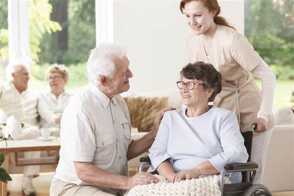 Starsza kobieta siedzi na wózku inwalidzkim, który pacha pielęgniarka przy wózku klęczy starszy mężczyzna. Seniorzy uśmiechają się do siebie