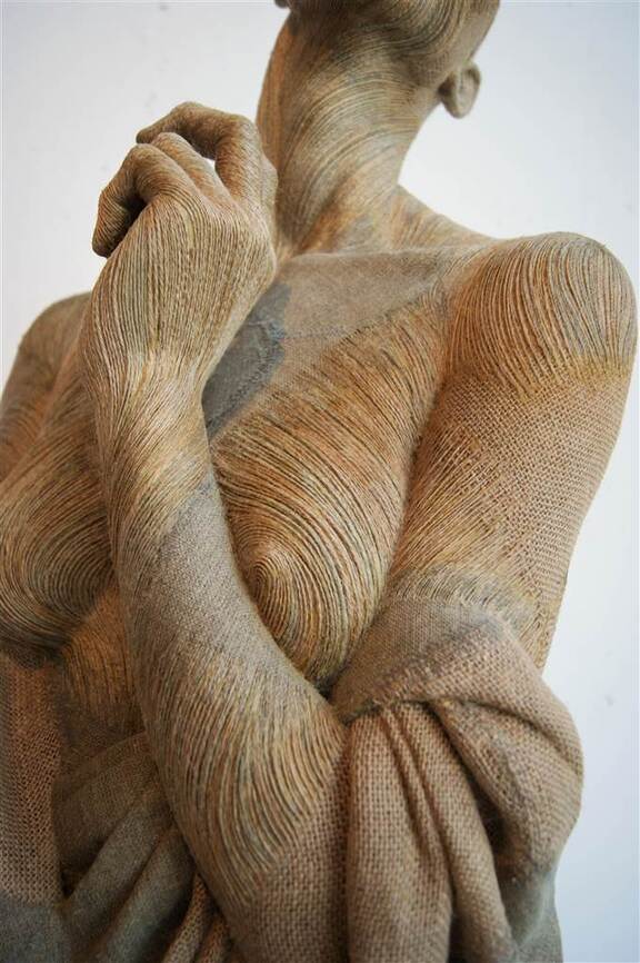 Zdjęcie przedstawia fragment rzeźby, prezentującej kobietę od szyi do pasa, z uniesioną do twarzy ręką i odsłoniętą piersią.
