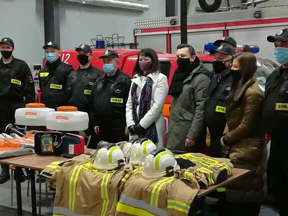 Członek zarządu Janina Ewa Orzełowska stoi wśród strażaków. Przed nimi na stole rozłozone są elementy stroju strażackiego oraz podarowny sprzęt