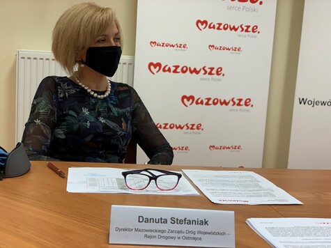 Dyrektor Danuta Stefaniak siedzi przy stole konferencyjnym lekko bokiem. Łokcie opiera o poręcze krzesła. Przed nią na stole jest dokument, a na nim rozłozone okulary. Dyrektor Stefaniak patrzy lekko w bok, jest w maseczce