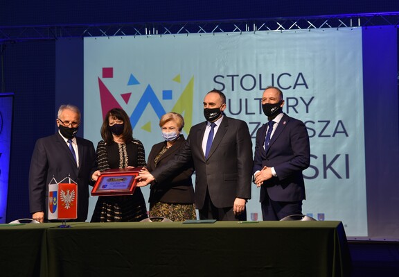 Elżbieta Lanc i Janina Ewa Orzełowska wraz z trzema przedstawicielami powiatu stoją za stołem i wspołnie trzymają klucz do stolicy kultury Mazowsza