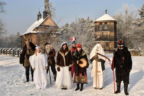 Grupa kolędników w przebraniach na tle zabudowań skansenu w Sierpcu. Zdjęcie w zimowej scenerii.