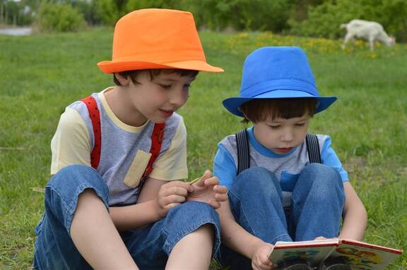 Dwóch chłopców w letnich kapeluszach siedzących na łące i czytających książkę.