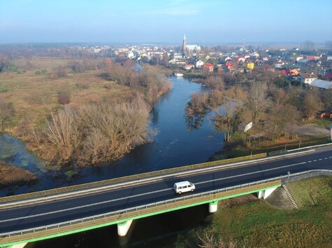 W dużym planie widok miejscowości, w oddali wiele budynków i płynąca rzeka - na pierwszym planie wiadukt na którym jedzie samochód osobowy.