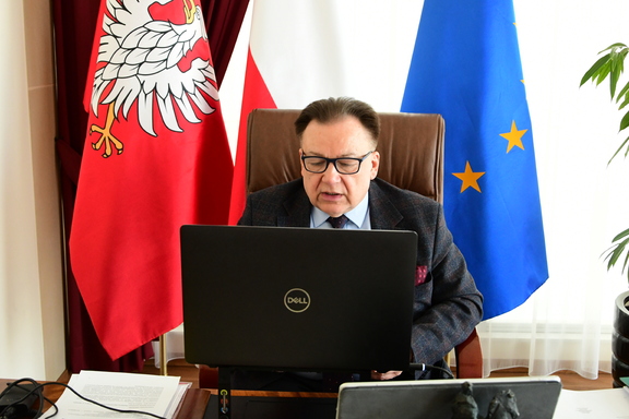 Marszałęk podczas zdalnej sesji, siedzi przy biurku przed otwartym laptopem, z tyłu stoją flagi UE, Polski i Mazowsza