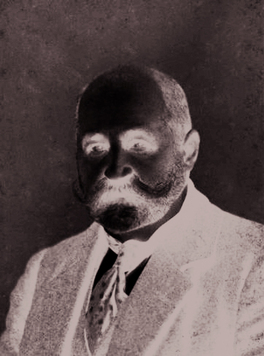 Łysiejący mężczyzna z bujnymi wąsami i starannie przystrzyżoną brodą. Ubrany w garnitur z kamizelką, kontrastowa koszula, pod szyją krawat.