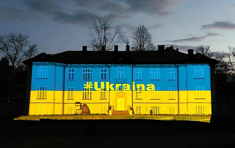 Fasada Pałacu Zespołu „Mazowsze” w pełnym planie w otoczeniu parkowej przyrody – oświetlona w barwy ukraińskie niebiesko-żółte z napisem Ukraina.