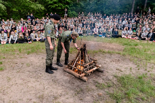 Dwóch harcerzy rozpala ognisko, wokół którego siedzi grupa haecerzy