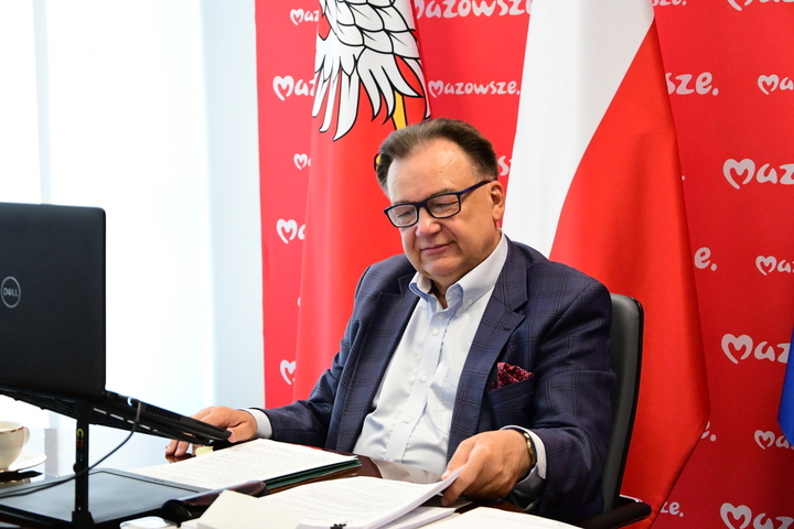 mężczyzna w garniturze siedzi przy biurku, na którym są dokumenty, w tle flaga Polski i Mazowsza
