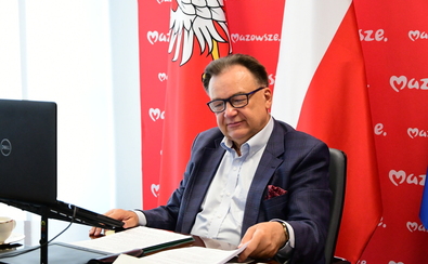 mężczyzna w garniturze siedzi przy biurku, na którym są dokumenty, w tle flaga Polski i Mazowsza