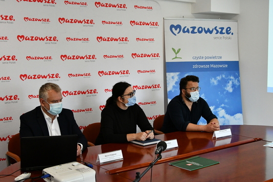 Troje osób za stołem, w tle ścianka promocyjna Mazowsza i rollup programu ochrony powietrza.