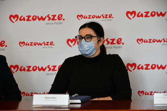 Burmistrzyni Kobyłki w okularach i maseczce siedzi przy stole, w tle ścianka Mazowsza.