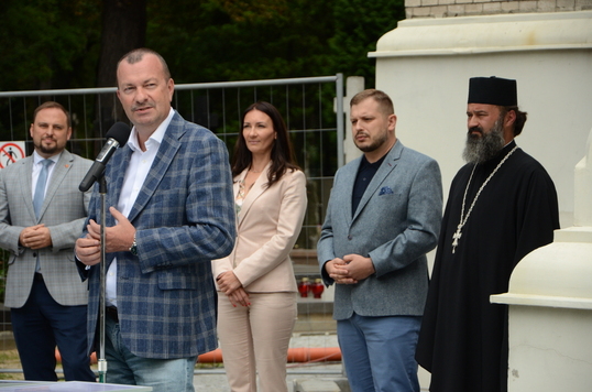 Elegancko ubrany mężczyzna mówi do mikrofonu, za nim grupa ludzi, wśród nich prawosławny duchowny