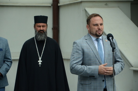 Elegancko ubrany mężczyzna mówi do mikrofonu, za nim stoi prawosławny duchowny.