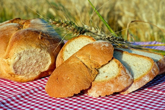Pokrojone kromki chleba ułożone nierówno na sobie na kraciastym obrusie. Na kromkach leżą kłosy zbóż.
