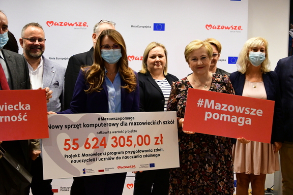 Elżbieta Lanc oraz Renata Kaznowska z beneficjnetami pozują do zdjęcia. Pani wiceprezydent trzyma symboliczny czek z kwotą ogólną przeznaczoną na program, a Elżbieta Lanc trzyma tablicę z napisem Mazowsze pomaga