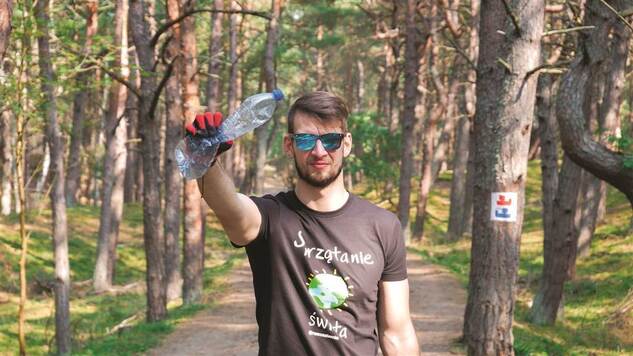 w lesie młody mężczyzna pozuje do zdjęcia, w dłoni trzyma plastikową butelkę