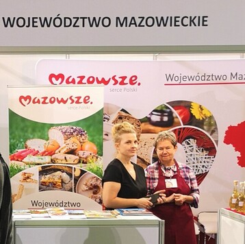 Dwie kobiety stoją na tle banerów reklamujących Mazowsze oraz mazowiecką zdrową żywność.