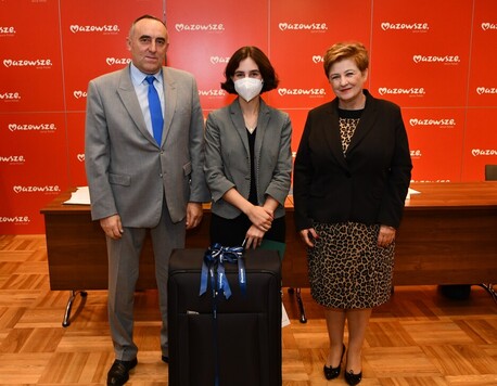 Młoda dziewczyna trzyma przed sobą walizkę, obok stoją kobieta i mężczyzna