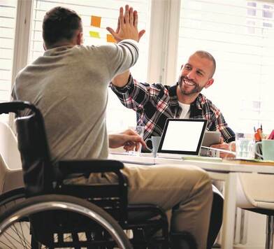 Mężczyzna na wózku inwalidzkim przybija dłoń uśmiechniętemu mężczyźnie siedzącemu przy biurku.