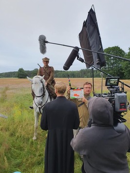 plan filmowy w plenerze, mężczyzna w mundurze wojskowym siedzi na białym koniu, wokół niego stoją aktorzy , kamerzysta i oświetleniowiec
