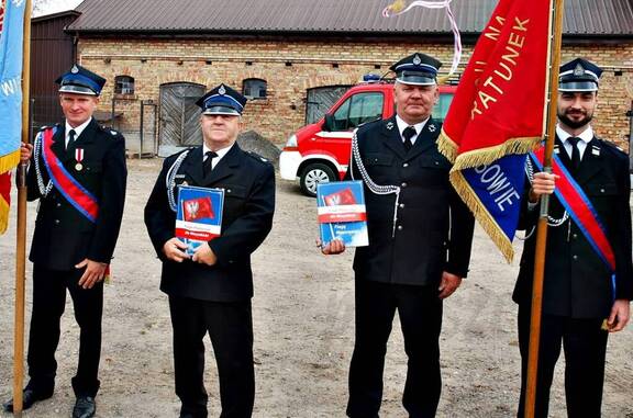 Czterech strażaków w oficjalnych mundurach stoi obok siebie. Przed sobą trzymają teczki z flagą Mazowsza. Jeden z nich trzyma sztandar