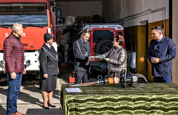 Kobieta przekazuje strażakowi teczkę aktową z dokumentami. Przed nimi na stole leżą nożyce hydrauliczne