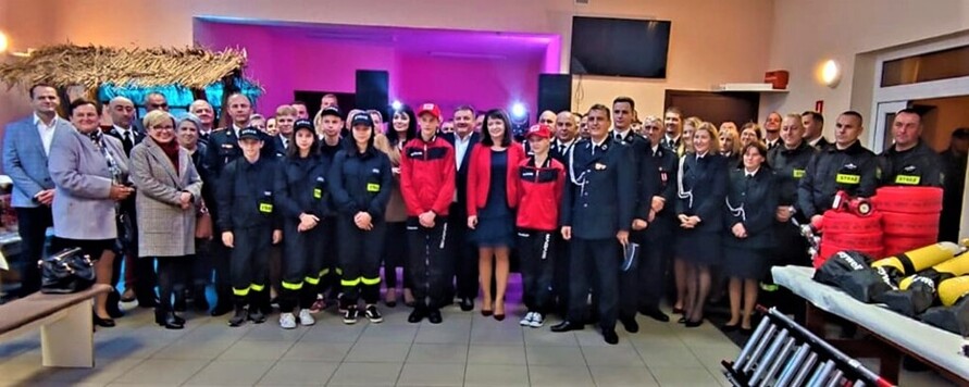 Grupa ludzi w strażackich mundurach, pośrodku kobieta w czerwonym żakiecie