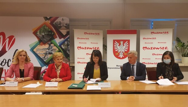 Elżbieta Lanc i Janina Ewa Orzełowska siedzą przy stole konferencyjnym i podpisują umowy. Obok nich siedzi radna joanna Bala i Beneficjenci