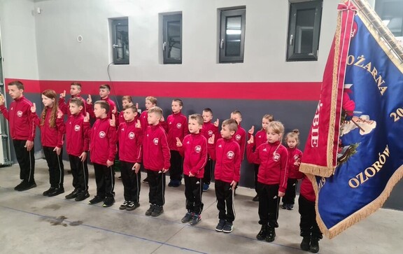 Dzieci ubrane w takie same mundurki stoją w dwóch rzędach i unoszą do góry prawą rękę.