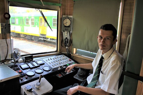 Kolejarz w kabinie maszynowej pociągu. Patrzy się prosto w kamerę, przed nim jest panel sterujący pociągu