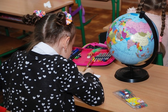 Dziewczynka z kucykami siedzi przy stoliku szkolnym i pisze w zeszycie. Przed nią stoi globus, a obok otworzony piórnik z kredkami