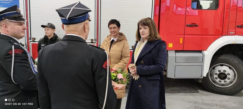 Janina Ewa Orzełowska stoi obok wójt gminy Stanisławów. Naprzeciw niej stoją strażacy, z którymi rozmawia