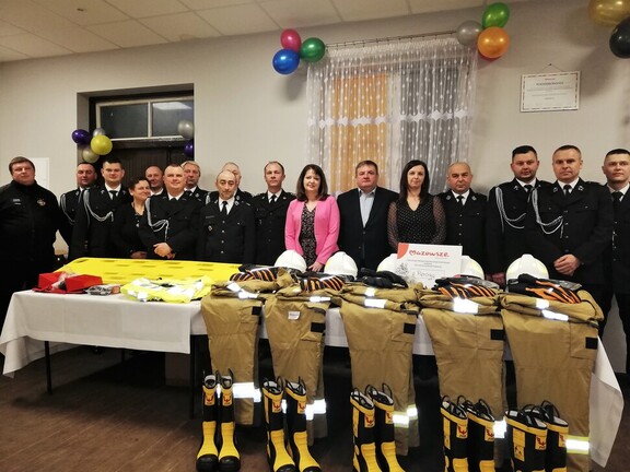Janina Ewa Orzełowska stoi wraz z grupą strażaków za stołem, na którym rozłożone są komplety odzieży ochronnej