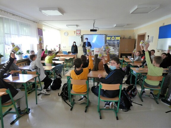 Uczniowie siedzą w szkolnych ławkach, w podniesionych prawych dłoniach pokazują odblaski. Przy tablicy stoi prowadzący zajęcia