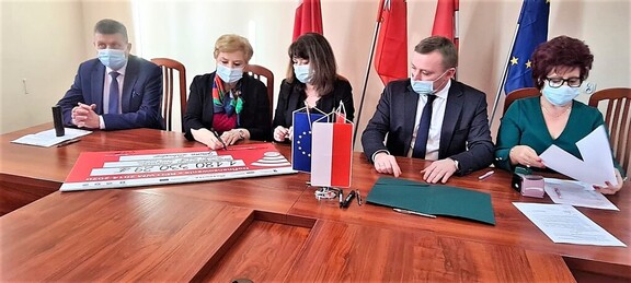 Janina Ewa Orzełowska, Elżbieta Lanc, wójt i skarbnik gminy siedzą przy stole i podpisują dokumenty oraz czek symboliczny