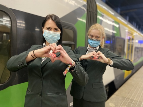 dwie kobiety stoją obok siebie, ręce mają ułożone w geście serca, w tle pociąg w zielono-biało-żółtych barwach Kolei Mazowieckich