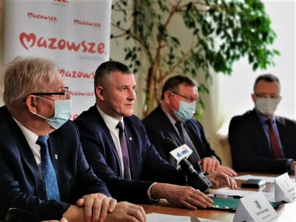 Wicemarszałek Rajkowski siedzi przy stole konferencyjnym i mówi do mikrofonu ustawionego na stole. Obok niego siedzi radny i dwóch przedstawicieli powiatu