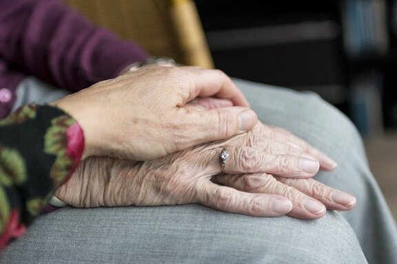 Dwie dłonie starszej osoby, złożone razem na kolanie. Trzecia dłoń przytrzymuje je w geście pomocy i opieki