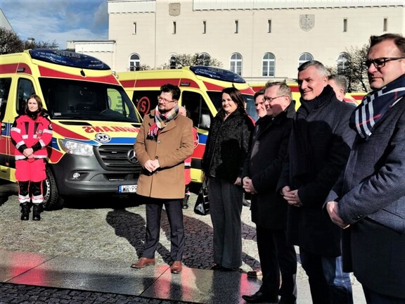 Wicemarszałek Rajkowski, radny Przybytniak oraz przedstawiciele lokalnych władz stoją obok siebie na placu. Za nimi widać nowe ambulanse