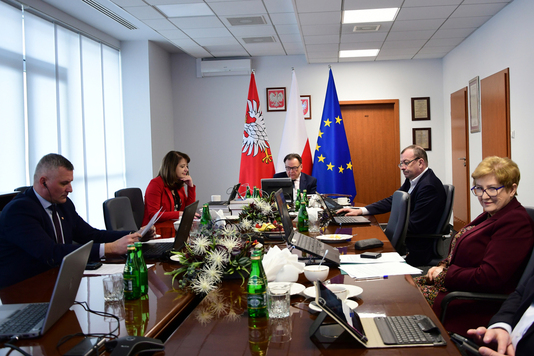 Grupa osób siedzi przy stole, w tle flagi: Mazowsza, Polski, Unii Europejskiej