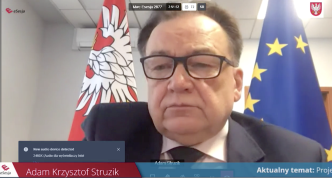 Stopklatka z internetowej transmisji sesji. Twarz marszałka w pełnym planie, za nim flaga Mazowsza i flaga Unii Europejskiej.