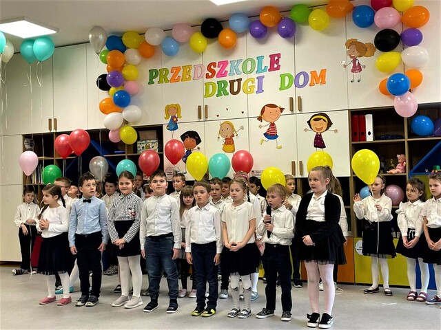 Grupa przedszkolaków stoi w sali zabaw przed widownią. Nad nimi, przy samym suficie, unoszą się baloniki