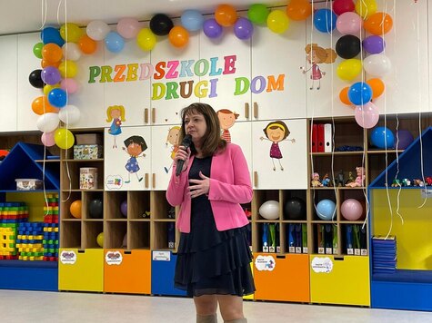 Janina Ewa Orzełowska stoi w sali przedszkolnej i mówi do mikrofonu, który trzyma w ręce