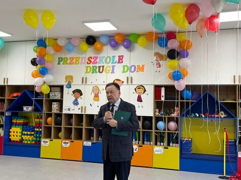 Marszałek stoi w przedszkolnej sali i mówi do mikrofonu, który trzyma w ręce