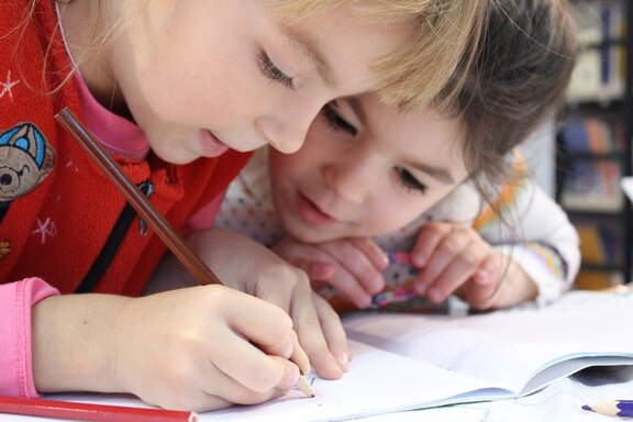 Dwoje dzieci w wieku szkolnym pochyla się nad zeszytem i wspólnie pracują. Trzymają w rękach ołówki.