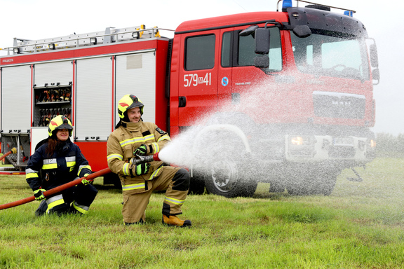 Dwóch strażaków (kobieta i mężczyzna) trzymają sikawkę, z której leci woda. W tle jest wóz strażacki.
